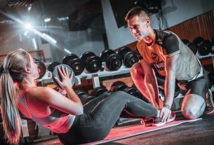 A personal trainer assists a woman doing sit-ups with a medicine ball in a gym setting.